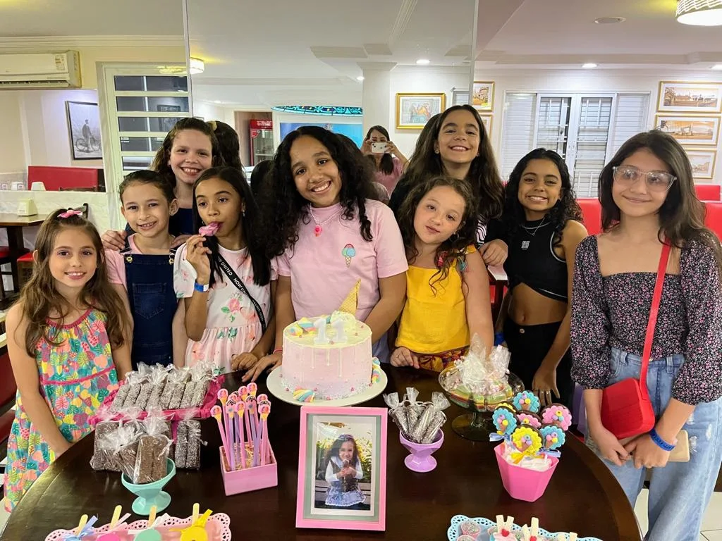 Aniversariante e as amigas em volta da mesa do bolo, com o salão ao fundo